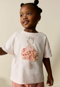 Tricou roz deschis din bumbac, cu o ilustrație florală a unei fete într-o rochie, având flori texturate din țesătură în nuanțe de piersic și roz.