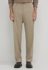 Pantaloni beige su misura con fronte piatto, tasche laterali e passanti per cintura, abbinati a scarpe nere su uno sfondo neutro.