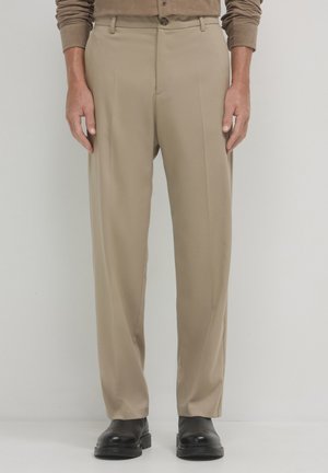 Pantalons beiges sur mesure avec une taille plate, poches latérales et passants de ceinture, associés à des chaussures noires sur un fond neutre.