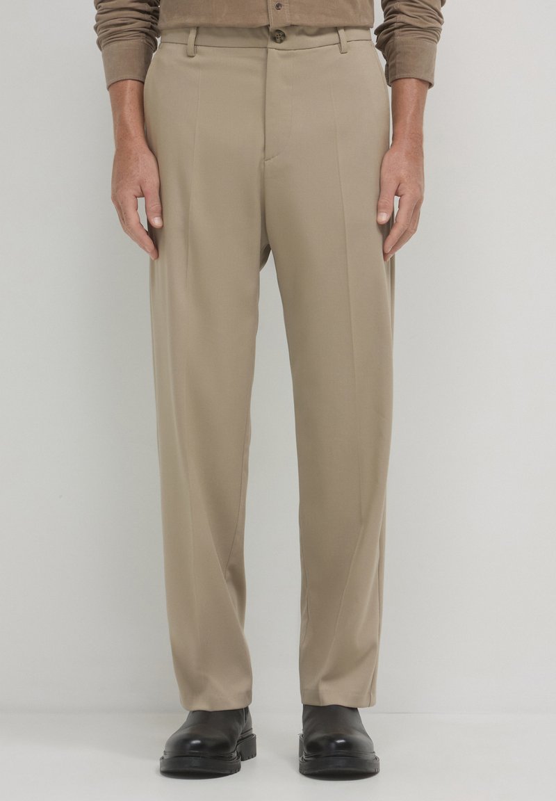 Pantaloni beige su misura con fronte piatto, tasche laterali e passanti per cintura, abbinati a scarpe nere su uno sfondo neutro.