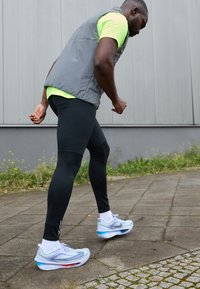 Sportchuhe mit einem hellblauen Obermaterial, das aus Mesh und Überzügen besteht, entworfen für das Laufen. Schwarze Leggings und eine graue Weste vervollständigen das Outfit.