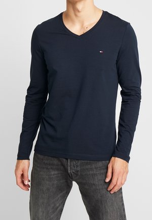 Mand iført en mørkeblå langærmet V-hals skjorte med lille logo på brystet og falmede sorte jeans, stående mod en ensfarvet baggrund.
