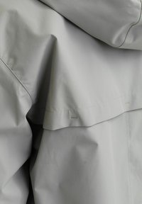 Šviesiai pilkas švarkas su lygiu, matinio paviršiaus tekstūru. Pasižymi sluoksniuota nugaros detale ir minimaliu siuvimu, kuris pabrėžia švarų dizainą.