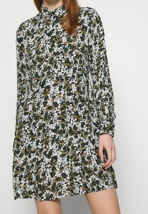 Vrouw draagt een minikleed met lange mouwen in een groen-wit-bruin bloemenpatroon en een kraag aan de hals.