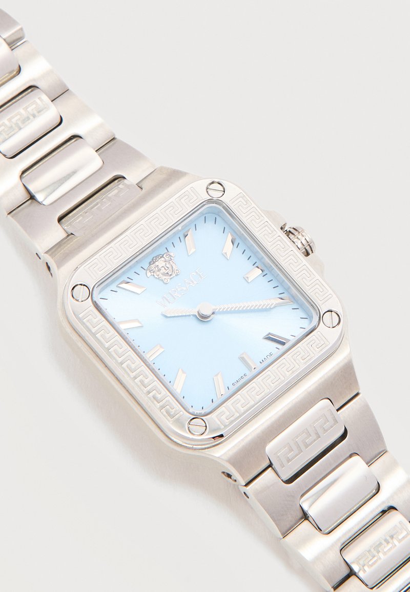 Silver Versace wristwatch with light blue square face, silver hour markers, Medusa logo, and Greek key pattern on bezel and band links.