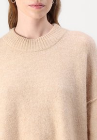 Beige genser med ribbestrikket rund hals, myk tekstur og avslappet passform, laget av lett materiale med en subtil glans.