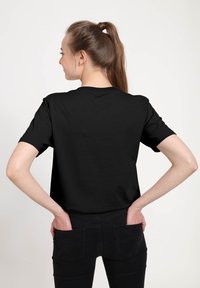 Černé tričko s krátkými rukávy vyrobené z měkké tkaniny, s kulatým výstřihem. Model má ruce v kapsách vzadu a na sobě má rovné černé džíny.