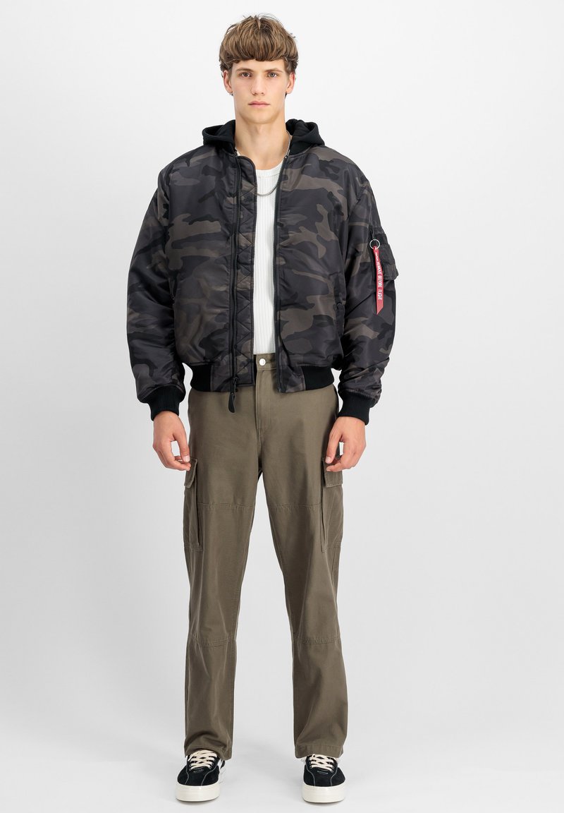 Giubbotto bomber camouflage con cappuccio, realizzato in nylon, dotato di zip frontale, polsini a coste e un'etichetta rossa sulla manica. Abbinato a pantaloni cargo color olivo.