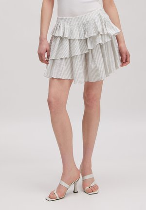 Vrouw draagt een wit en lichtgrijs gestreept gelaagd minirokje met witte sandaaltjes met hoge hakken en bandjes, staand op een effen achtergrond.