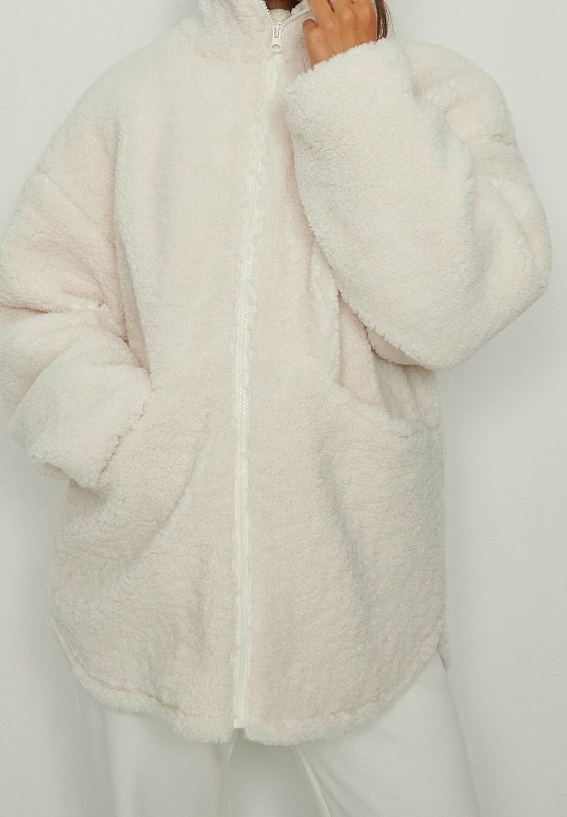 Persona que lleva una chaqueta de forro polar gruesa, de gran tamaño y color blanco roto, con bolsillos, sosteniendo una mano cerca de su cara.