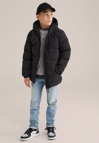 Juodas pūkinis švarkas su gobtuvu, užtrauktuku ir elastingomis rankovėmis, dėvimas ant pilkos džemperio. Šviesiai mėlynos džinsinės kelnės ir juodi sportbačiai užbaigia aprangą.