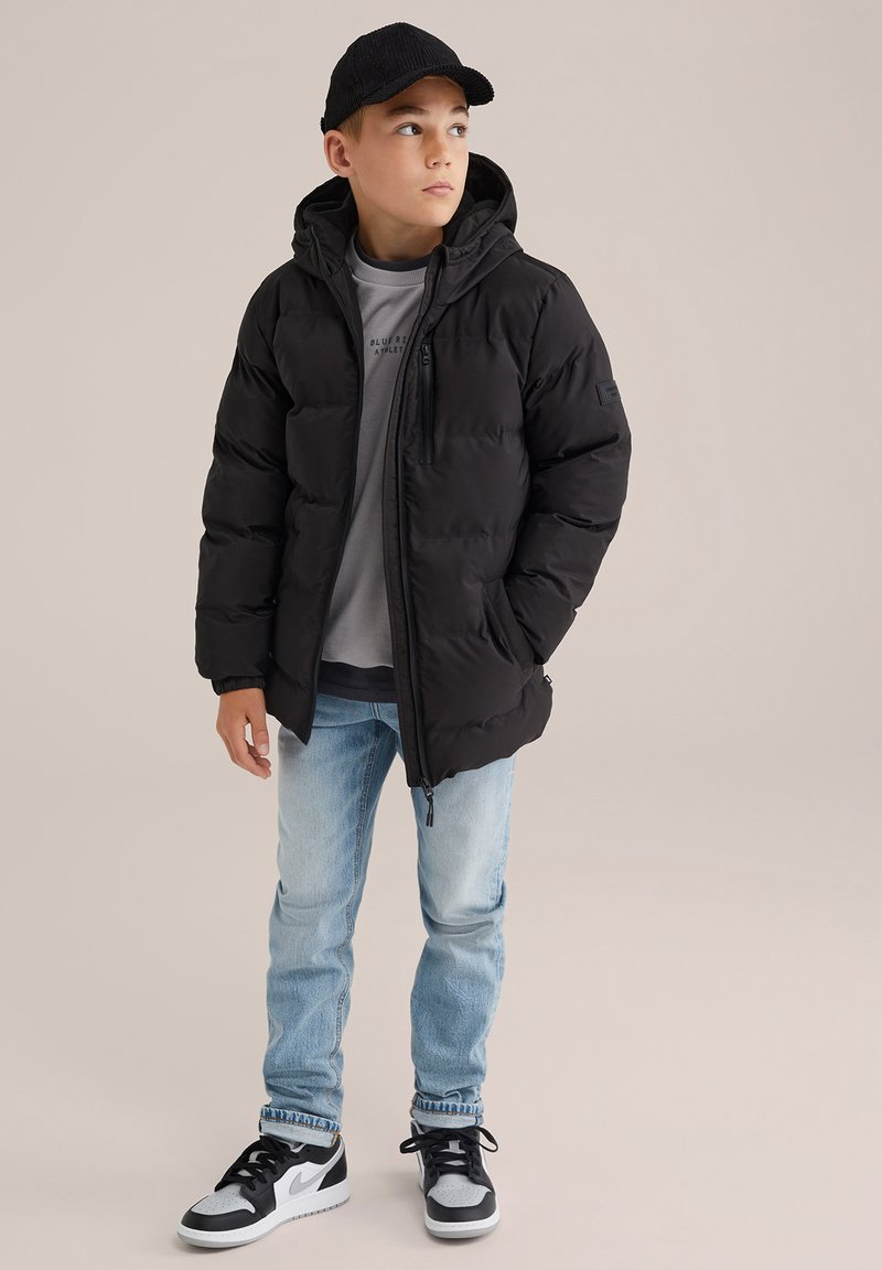 Juodas pūkinis švarkas su gobtuvu, užtrauktuku ir elastingomis rankovėmis, dėvimas ant pilkos džemperio. Šviesiai mėlynos džinsinės kelnės ir juodi sportbačiai užbaigia aprangą.