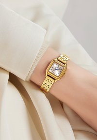 Montre-bracelet carrée en or avec un cadran blanc, des index horaires noirs et une aiguille des secondes bleue. Bracelet en maillons métalliques avec finition brillante.