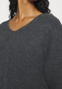 Pull gris foncé en maille avec un col en V, présentant une finition douce et texturée ainsi que des motifs de côtes diagonales prononcés.