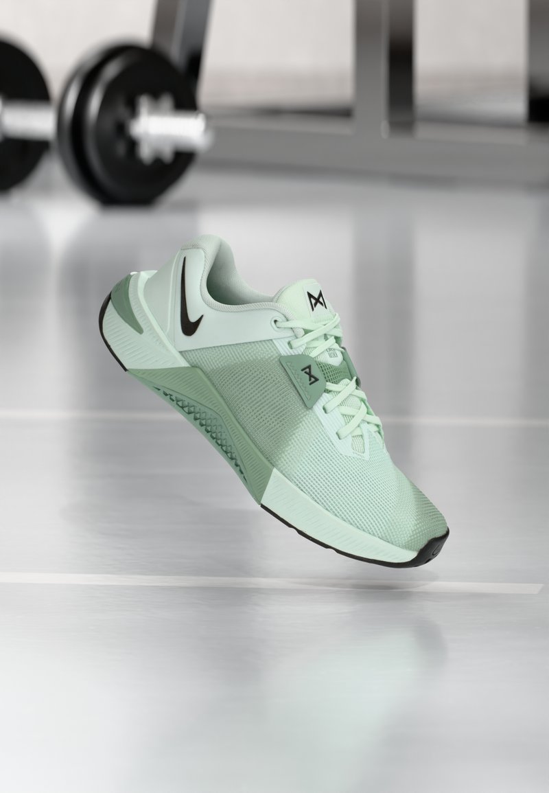 Chaussure de sport vert clair avec tige en maille, accents noirs, semelle texturée et lacets. Présente un logo signature sur la languette et le côté.