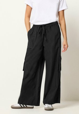 Femme portant un pantalon cargo noir à jambes larges avec taille à cordon, un t-shirt blanc et des baskets blanches avec des bandes noires.