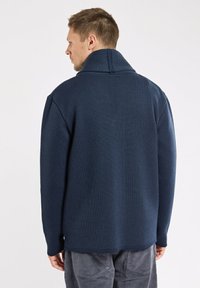 Homme de dos portant un pull en laine texturé bleu marine avec un col haut et un pantalon en velours côtelé gris, devant un fond uni.