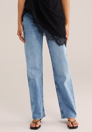 Naine, kes kannab helesiniseid sirge säärega pükse, musta pitsiga kaunistatud topi ja musti rihmikuid kontsakingi, seisab käed lõdvestunult.