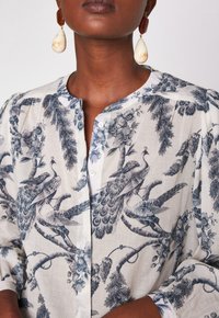 Donna che indossa una blusa bianca con stampa di pavone blu e fiori, abbinata a grandi orecchini a goccia color crema.