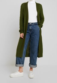 Cardigan en maille vert olive avec design à ouverture avant, porté sur un col roulé blanc et un jean taille haute bleu clair, assorti à des baskets blanches.
