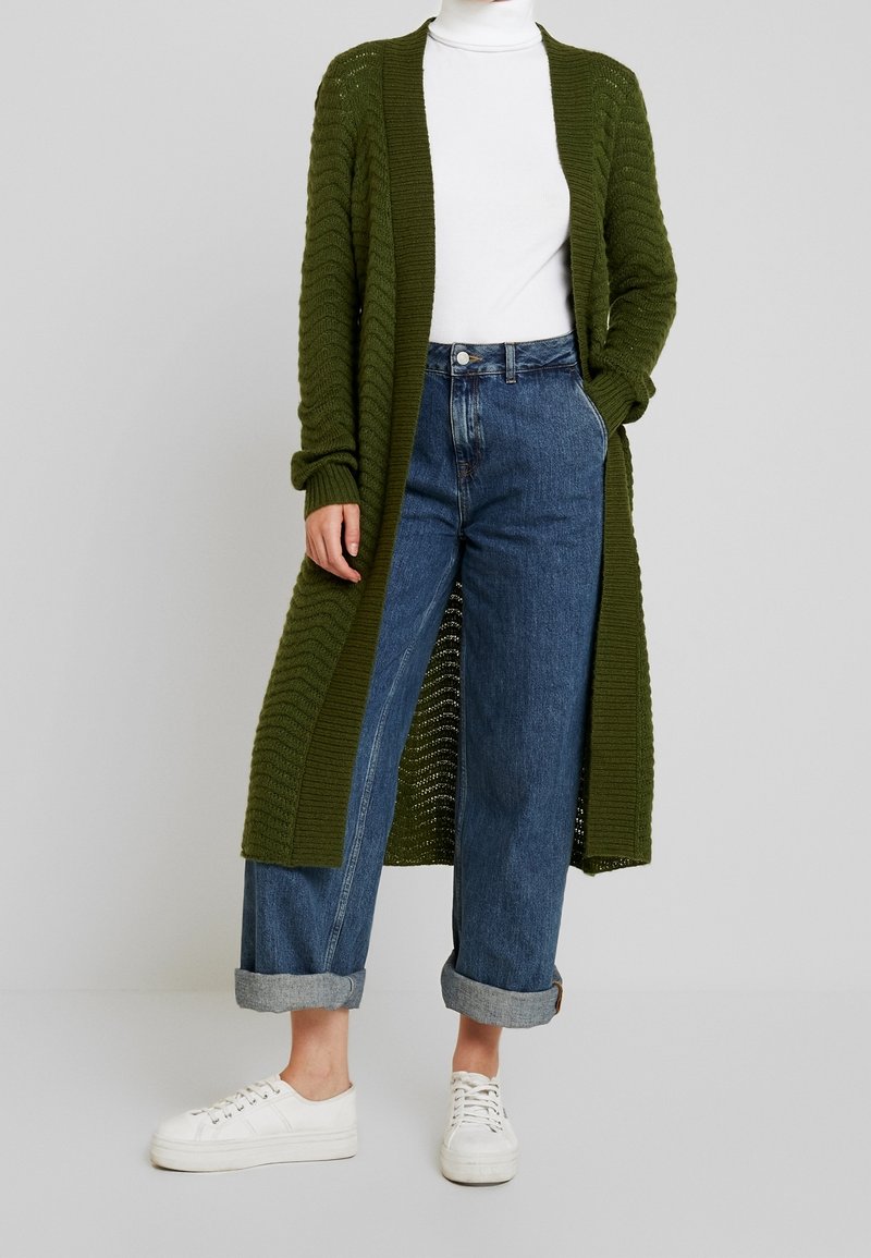 Cardigan en maille vert olive avec design à ouverture avant, porté sur un col roulé blanc et un jean taille haute bleu clair, assorti à des baskets blanches.
