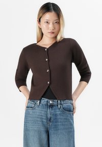 Vrouw met schouderlang blond haar, gekleed in een bruine knopencardigan en losse blauwe spijkerbroek, staand met haar handen in haar zakken.