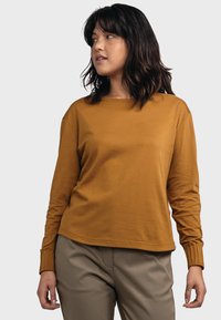 Langärmliges, senfgelbes Baumwollhemd mit rundem Halsausschnitt und gerippten Bündchen, kombiniert mit hellen Hosen. Sanfte Textur und lässige Passform.