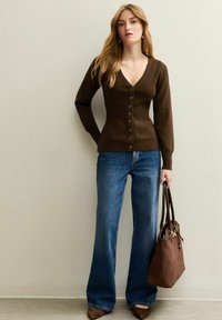 Donna che indossa un cardigan marrone con bottoni e scollo a V, jeans a gamba larga blu, scarpe a punta marroni, mentre tiene una borsa marrone in mano, in piedi contro un muro beige.