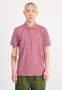 Bavlnená polo košeľa v tlmených ružových farbách s troj-buttonovou légou, krátkymi rukávmi a malým žltým logom na hrudi. Detail textúrované tkaniny.