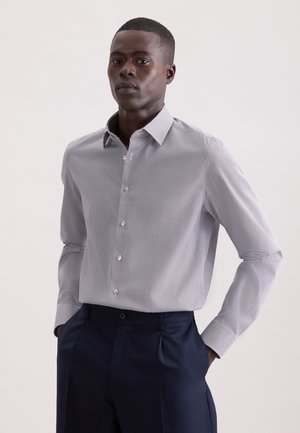 Homme portant une chemise ajustée à petits carreaux blancs et gris à manches longues et un pantalon habillé bleu marine, debout les mains dans les poches.