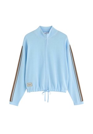 Hellblaue Jacke mit Reißverschluss, braunen Doppelstreifen an den Ärmeln, einem Kordelzug am Saum und einem kleinen Patch mit der Aufschrift „Social“ vorne links.
