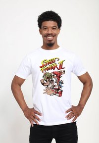 Bílý bavlněný tričko s barevným grafickým motivem "Street Fighter II" s postavami v akci. Model stojí sebevědomě, ruce na bocích.