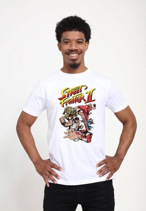 White cotton t-shirt featuring a colorful "Street Fighter II" graphic with characters in action. Model stands confidently, hands on hips.