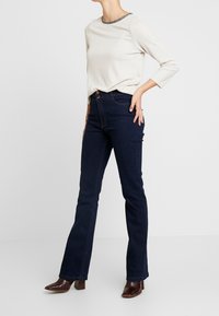 Un haut beige clair à manches longues avec une bordure grise décorative au niveau du col, associé à un jean évasé bleu foncé et des bottines à talons marron.