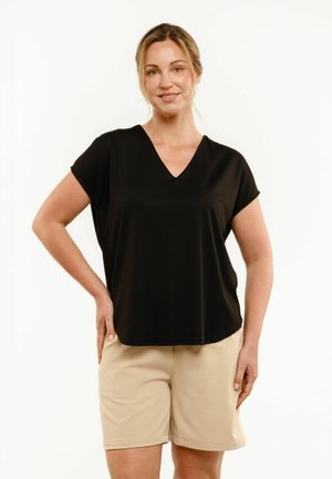 Femme debout, main sur la hanche, portant un haut noir à manches courtes col en V et un short beige arrivant aux genoux sur fond blanc.