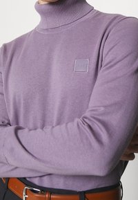 Person som bär en lavendelfärgad polotröja med en "BOSS" logotyp patch, med armarna i kors, svarta byxor och ett brunt läderbälte.