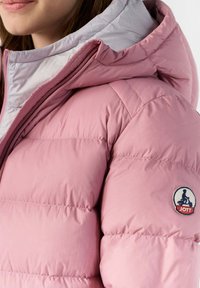 Doudoune rose avec une capuche gris clair contrastante, texture matelassée et un badge logo JOTT sur le côté gauche.