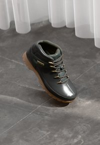 Bot de drumeție de culoare verde închis și negru, cu un finisaj lucios, accente texturate, elemente de bronz aurit și o talpă ușoară de culoare bej.