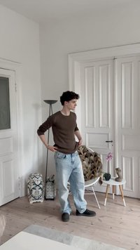 Jersey de canalé marrón claro combinado con jeans azul claro y zapatos negros. Paredes blancas, suelo de madera, silla decorativa, pequeña mesa con decoración.