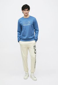 Blå sweatshirt med vitt grafiskt motiv, matchad med cremefärgade sweatpants med svart logotryck. Modellen bär vita sneakers och står upprätt.