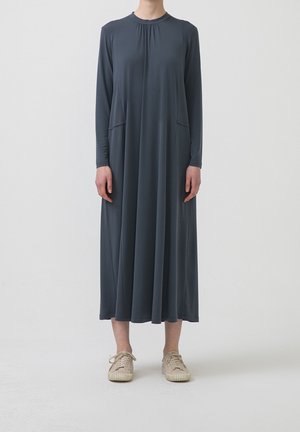 Osoba ubrana w ciemnoszarą, długą suknię z długim rękawem sięgającą do kostek z bocznymi kieszeniami i beżowymi trampkami, stojąca na prostym jasnym tle.