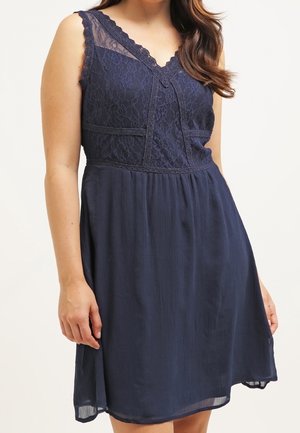 Ärmelloses dunkelblaues Kleid mit Spitzenoberteil, V-Ausschnitt und plissiertem Rock, getragen von einer Frau vor einem einfarbigen Hintergrund.