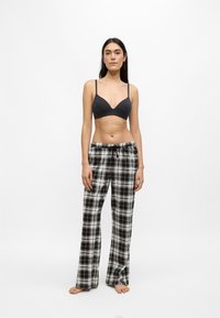 Kvinna som står barfota och bär en svart bralette och svartvita rutiga pyjamasbyxor mot en enkel vit bakgrund.