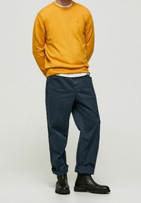 Gelber Strickpullover, dunkelblaue Hose und schwarze Stiefeletten. Der Pullover hat einen Rundhalsausschnitt und ein gesticktes Logo; die Hose sitzt etwas lockerer.