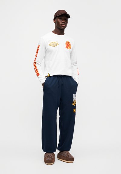 Λευκό πουκάμισο με μακριά μανίκια με κόκκινα και κίτρινα γραφικά, σε συνδυασμό με σκούρο μπλε παντελόνι φόρμας με κίτρινες λεπτομέρειες και καφέ καπέλο. Περιλαμβάνονται καφέ παντόφλες.