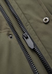 Gros plan d'une fermeture éclair noire et de boutons-pression métalliques sur un tissu vert, montrant les détails de la couture et le curseur de la fermeture avec le nom de la marque "ombre".