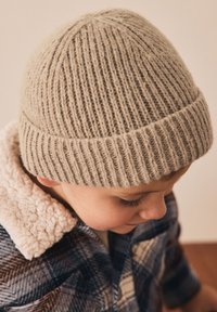 Bonnet en tricot côtelé brun clair avec un revers. Porté par un enfant, il présente une texture douce et un ajustement confortable, associé à une veste à carreaux.