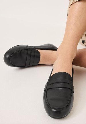Mocassins en cuir noir avec des bouts ronds, des détails cousus et une sangle à l'avant sur le coup de pied. Texture lisse avec un profil bas.