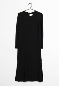 Robe noire à manches longues en tissu côtelé avec un col rond et des fentes latérales pour faciliter le mouvement. Suspendue à un crochet noir.
