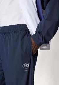 Mornarske športne hlače z elastičnim pasom, stranskimi žepi in belimi logotipnimi detajli. Predstavljena jakna ima bele in mornarsko modre sekcije ter zadrgo.